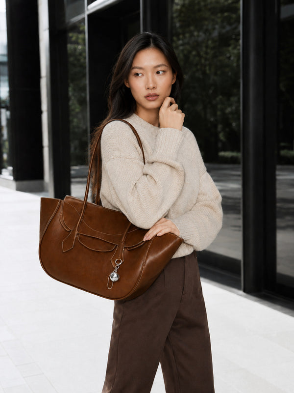 Large winged leather-look tote bag with single arched handle, stitched flap-front panel, and silver ball charm in cognac