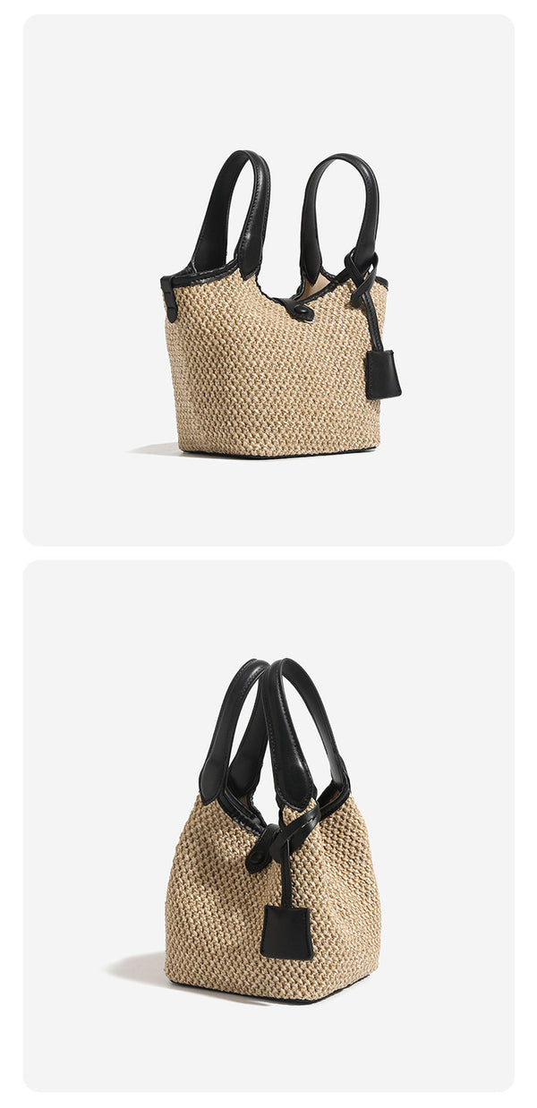 Woven beige straw bucket bag with black leather-look trim, dual top handles, detachable shoulder strap, and front snap