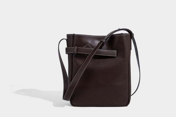 Structured dark brown leather-look bucket bag with white contrast stitching, front wraparound belt buckle strap, and long