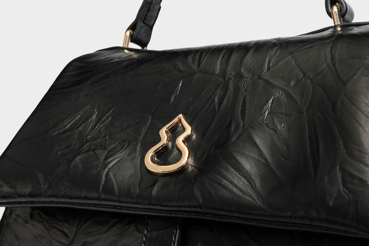 Structured black satchel bag with glossy crinkled leather-look finish, gold-tone emblem flap closure, slim top handle, and