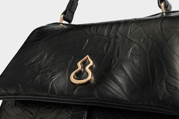 Structured black satchel bag with glossy crinkled leather-look finish, gold-tone emblem flap closure, slim top handle, and