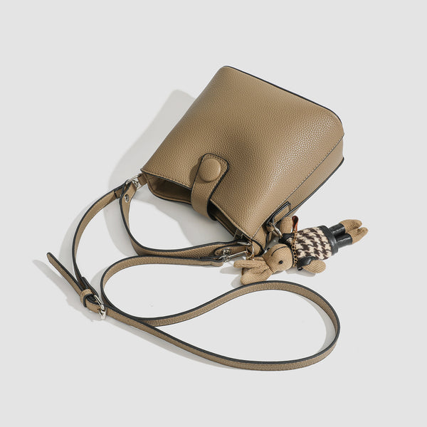 Structured taupe pebbled-texture bucket bag with top handle, silver ring hardware, tab-and-button closure, and knit bunny