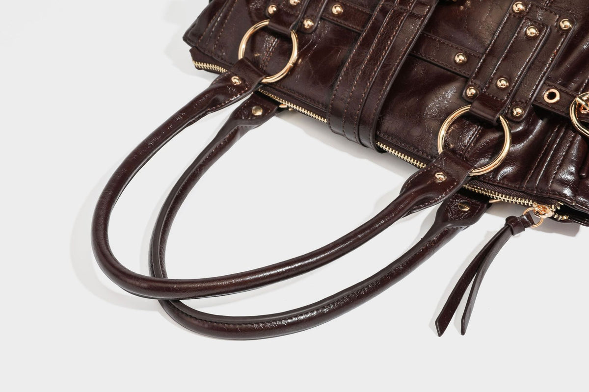 Glänzende Henkeltasche in Lederoptik mit zwei oberen Henkeln, goldfarbenen Nieten- und Ringbeschlägen, einem Riemendetail mit Schnalle auf der Vorderseite und