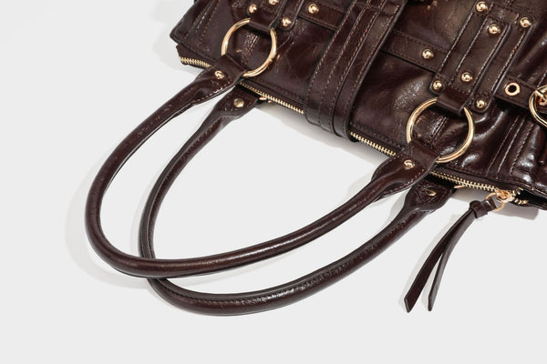 Glänzende Henkeltasche in Lederoptik mit zwei oberen Henkeln, goldfarbenen Nieten- und Ringbeschlägen, einem Riemendetail mit Schnalle auf der Vorderseite und
