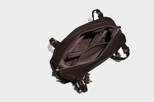 Weiche, zylinderförmige Schultertasche in Dunkelbraun mit silberfarbenen Riemen mit Ösen, Reißverschluss oben, seitlichen Schnallenbeschlägen und Quaste
