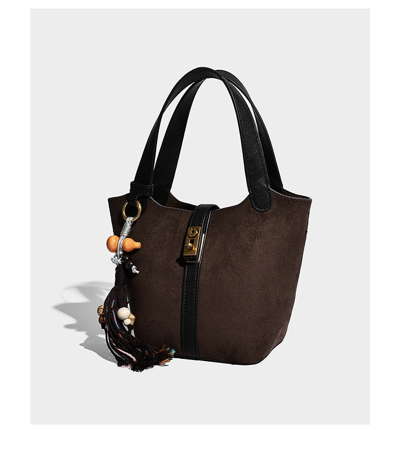 Small structured trapezoid tote bag in matte suede-look finish with black contrast trim, gold-tone front clasp, tassel charm