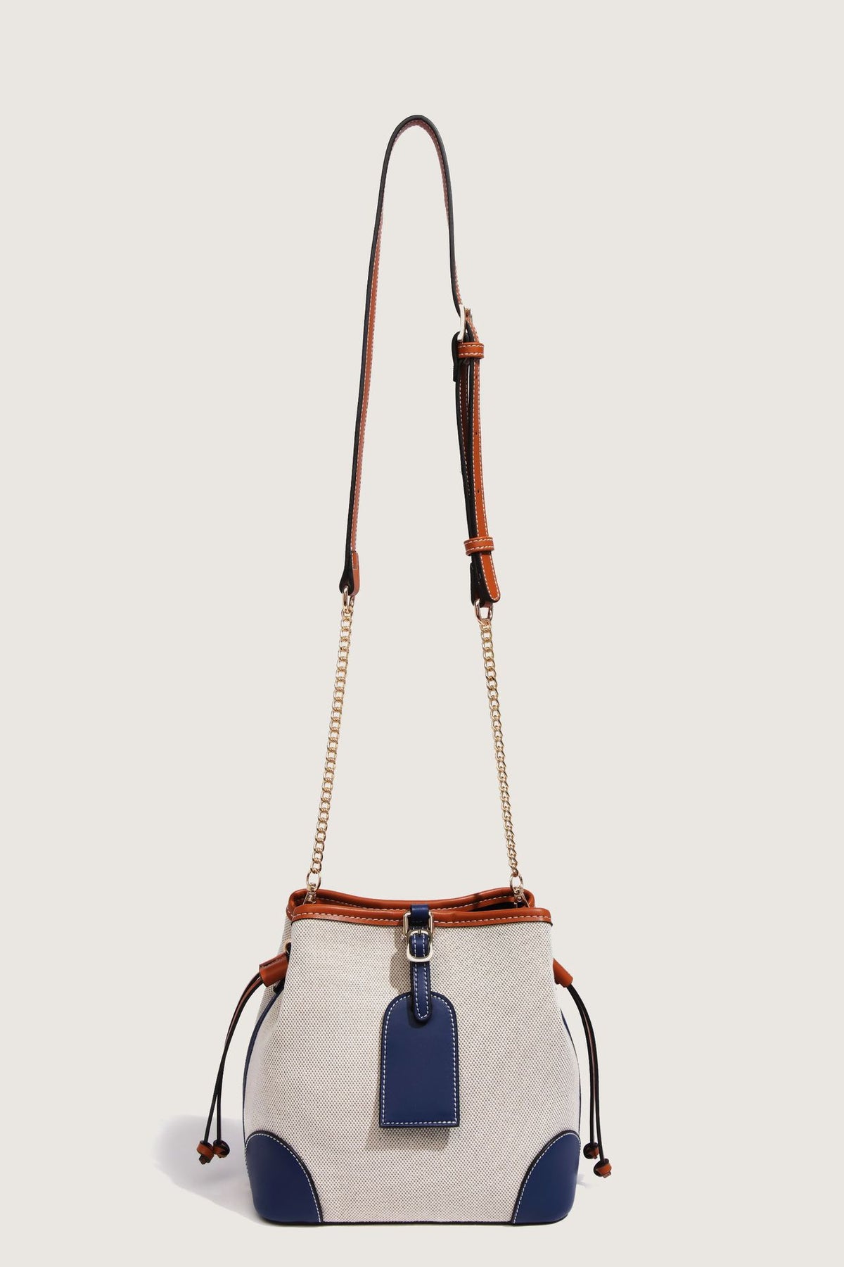 Cream woven-texture canvas bucket bag with navy accent panels, tan leather-look trim, gold-tone chain strap, and tassel