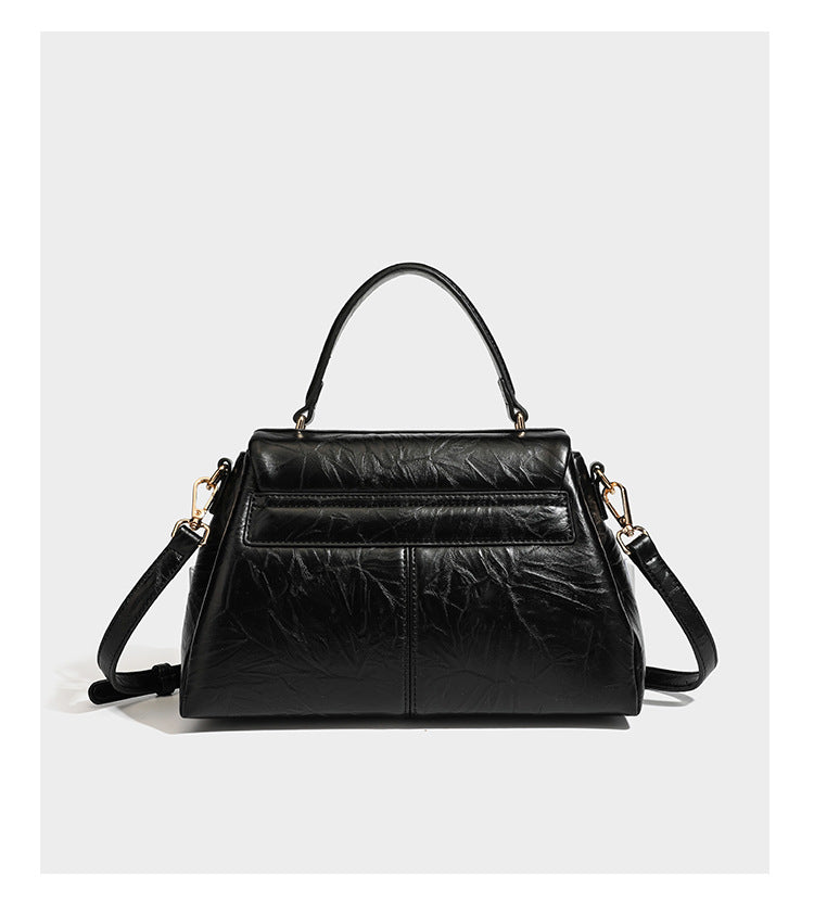 Structured black satchel bag with glossy crinkled leather-look finish, gold-tone emblem flap closure, slim top handle, and
