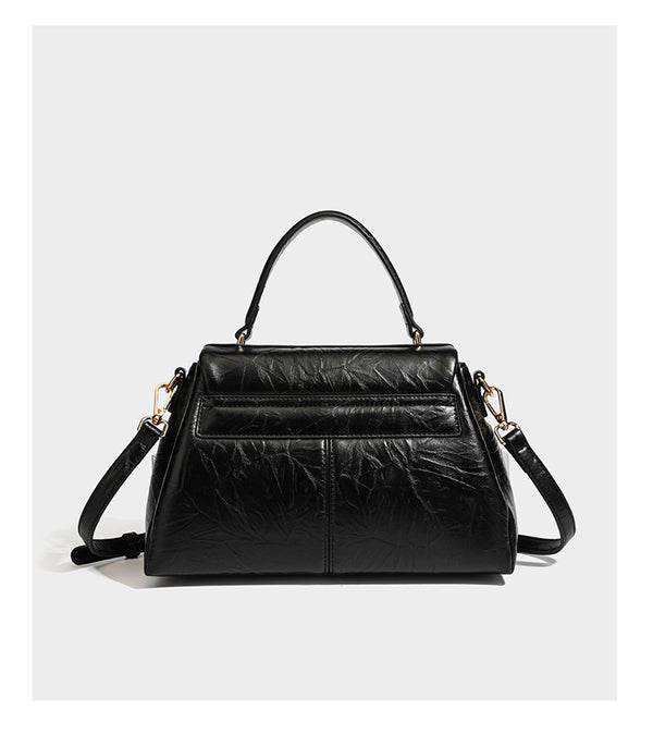 Structured black satchel bag with glossy crinkled leather-look finish, gold-tone emblem flap closure, slim top handle, and