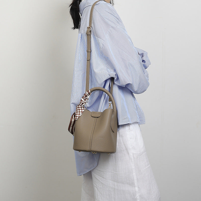Pebbled leather-look bucket bag with checkered scarf-wrapped top handle, flap tab closure, gold-tone hardware, and matching