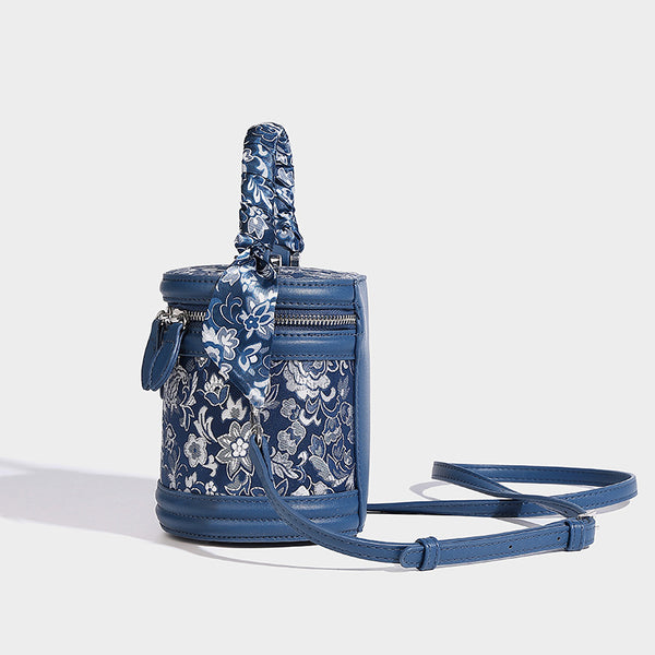 Small cylindrical barrel bag in blue-and-white floral jacquard with leather-look trim, scarf-wrapped top handle, silver-tone