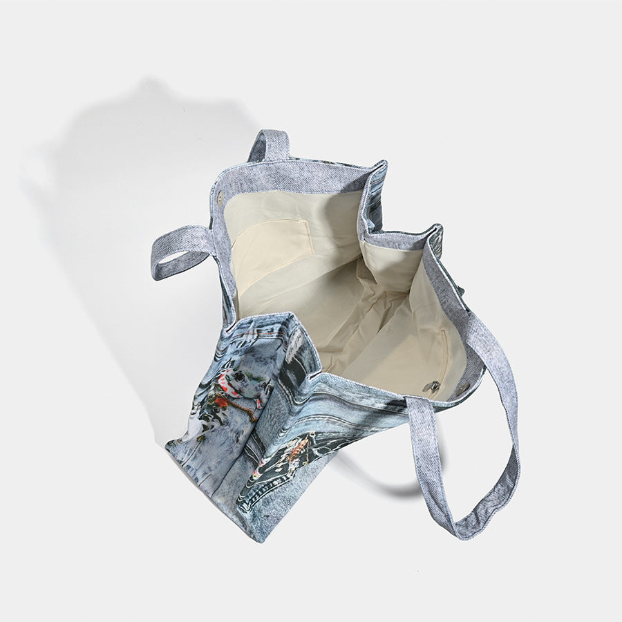 Large open-top tote bag with printed denim jeans graphic in faded blue-grey, dual flat shoulder straps and rivet hardware