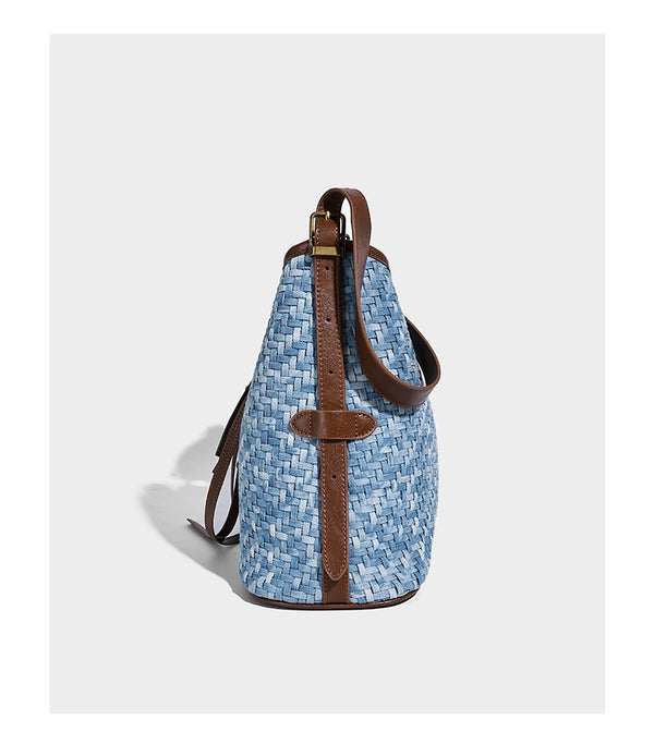 Light blue woven bucket bag with brown leather-look trim, gold-tone hardware, adjustable shoulder strap, and decorative bead