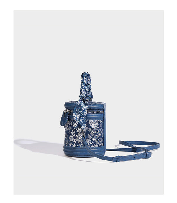 Small cylindrical barrel bag in blue-and-white floral jacquard with leather-look trim, scarf-wrapped top handle, silver-tone