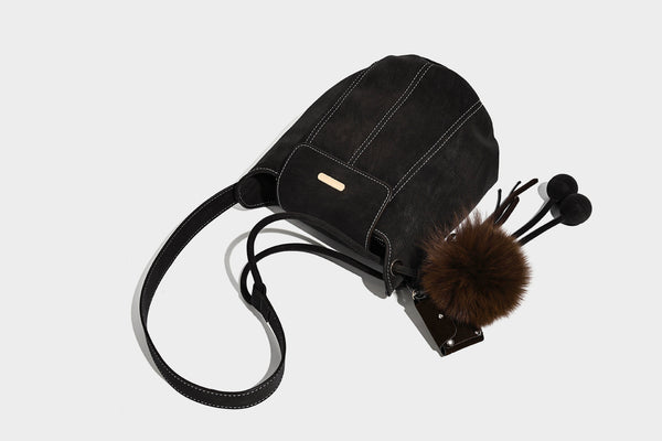 Small matte textured bucket crossbody bag with flap closure, gold-tone bar hardware, contrast stitching, and pompom tassel