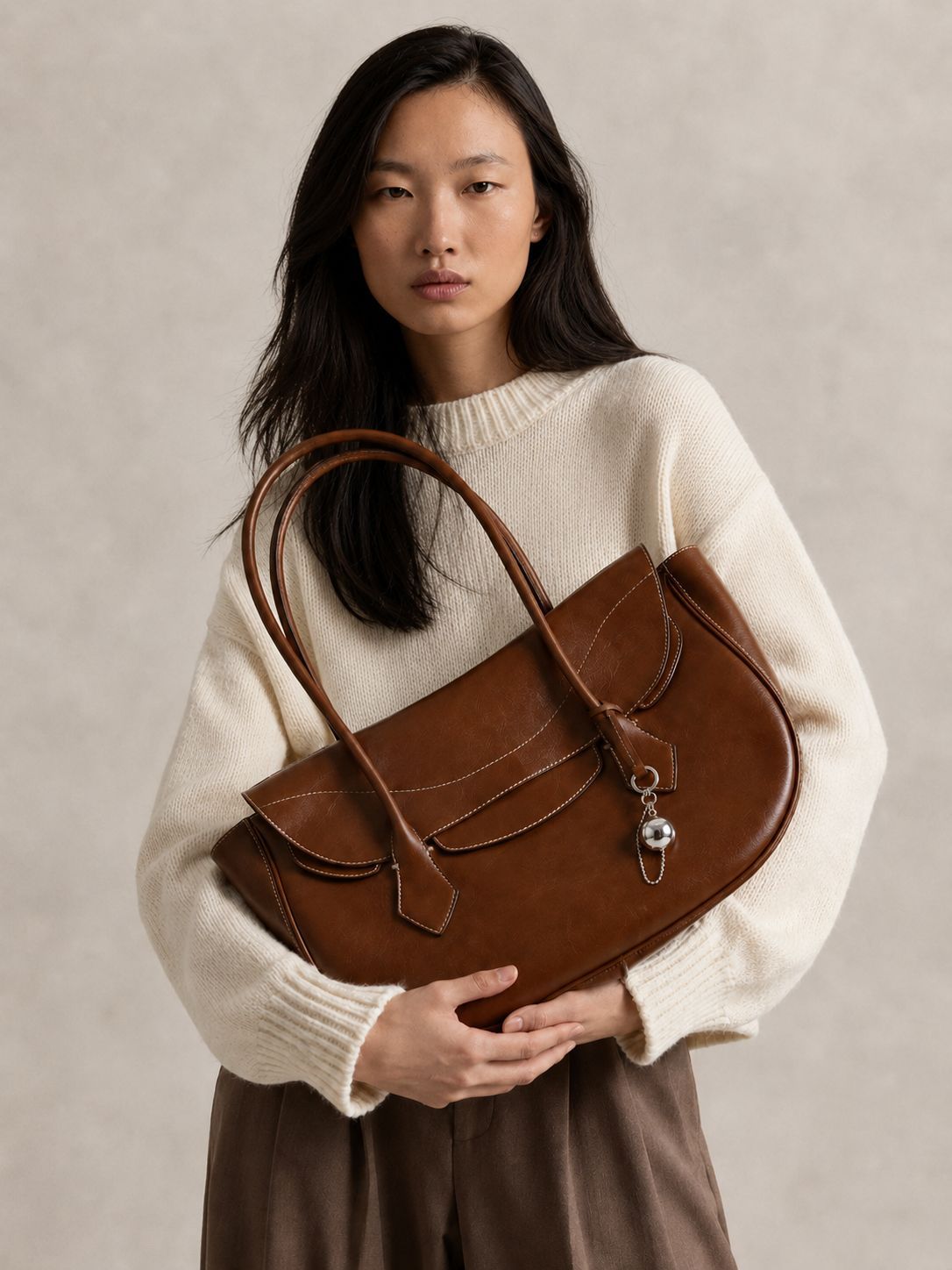 Large winged leather-look tote bag with single arched handle, stitched flap-front panel, and silver ball charm in cognac