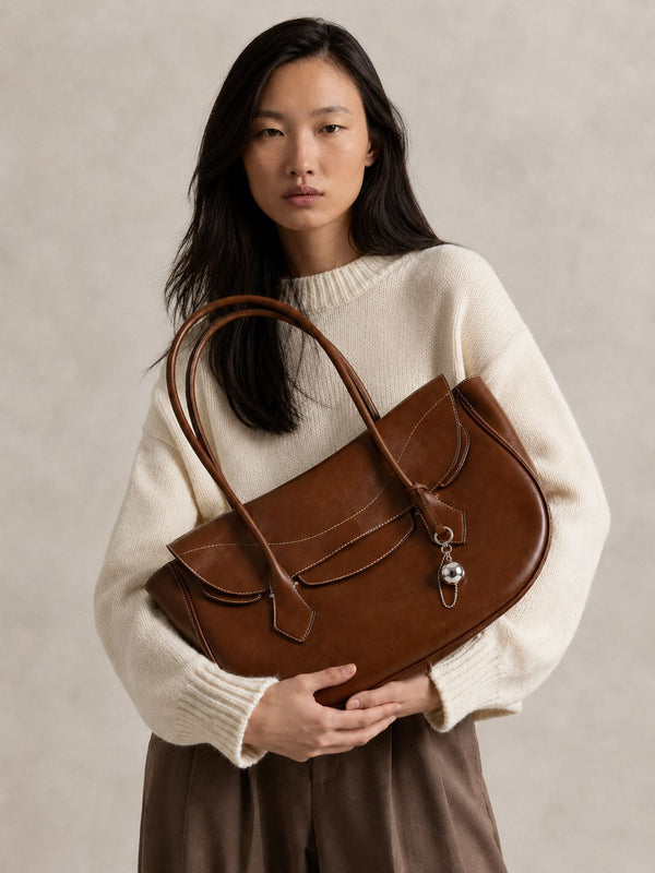 Large winged leather-look tote bag with single arched handle, stitched flap-front panel, and silver ball charm in cognac
