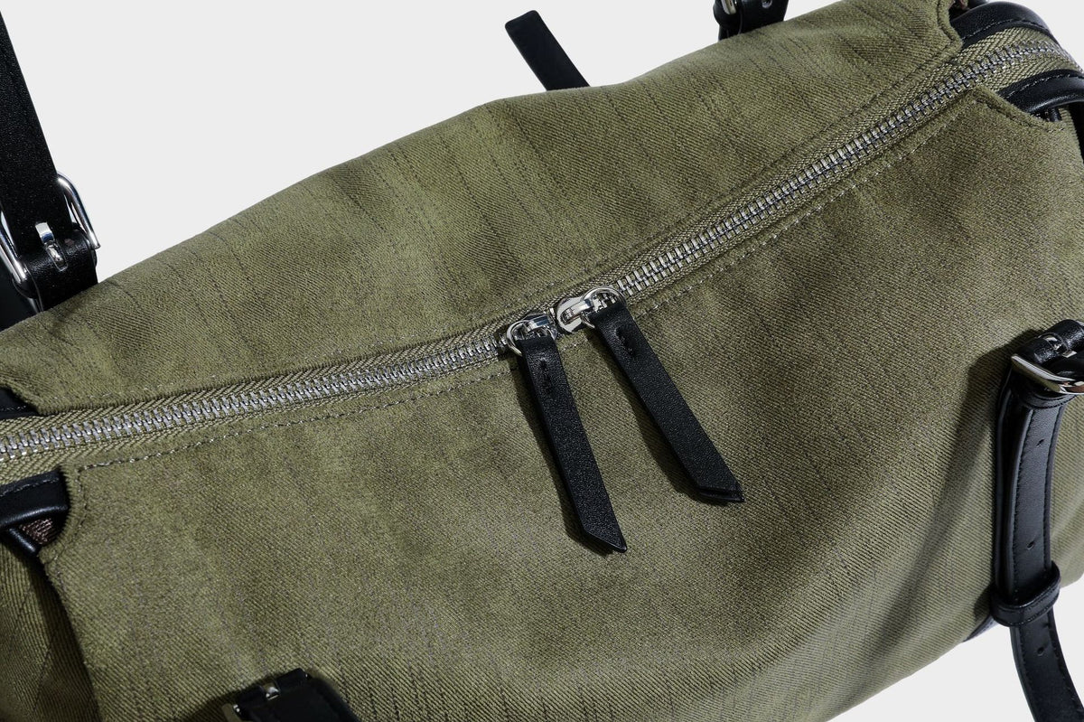 Olivgrüne Canvas-Schultertasche in Zylinderform mit schwarzen Trägern in Lederoptik, Reißverschluss oben und silberfarbenen Metallbeschlägen, Detailansicht