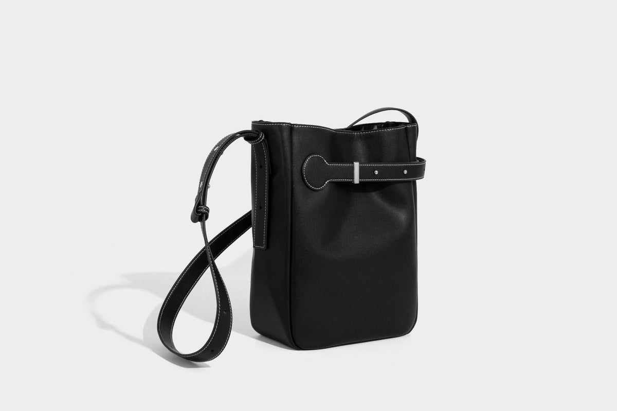 Structured dark brown leather-look bucket bag with white contrast stitching, front wraparound belt buckle strap, and long