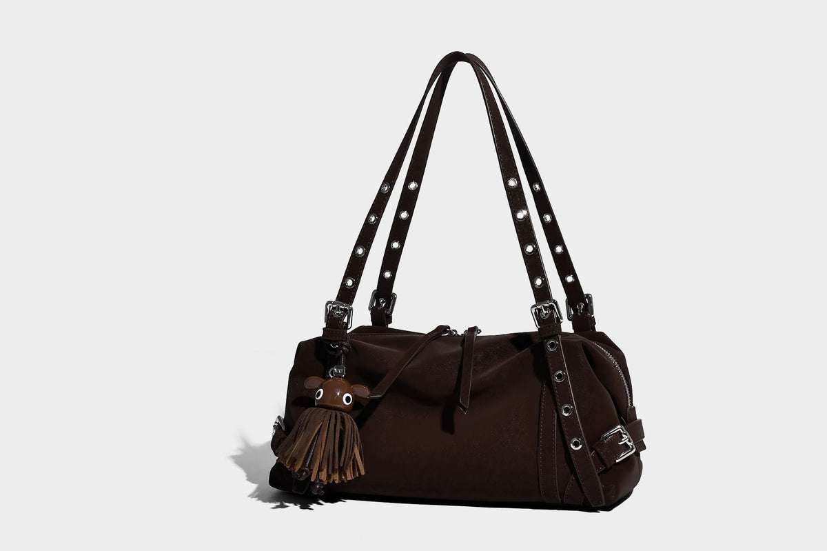 Weiche, zylinderförmige Schultertasche in Dunkelbraun mit silberfarbenen Riemen mit Ösen, Reißverschluss oben, seitlichen Schnallenbeschlägen und Quaste