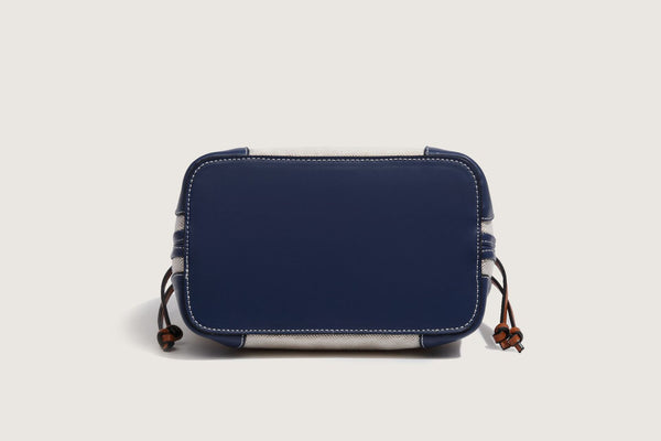 Cream woven-texture canvas bucket bag with navy accent panels, tan leather-look trim, gold-tone chain strap, and tassel