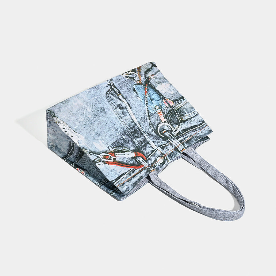 Large open-top tote bag with printed denim jeans graphic in faded blue-grey, dual flat shoulder straps and rivet hardware
