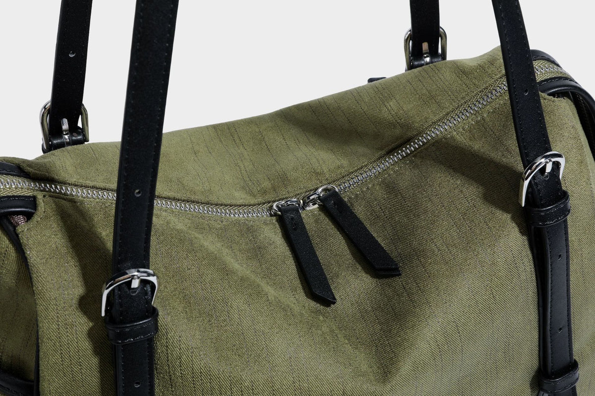 Olivgrüne Canvas-Schultertasche in Zylinderform mit schwarzen Trägern in Lederoptik, Reißverschluss oben und silberfarbenen Metallbeschlägen, Detailansicht