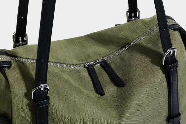 Olivgrüne Canvas-Schultertasche in Zylinderform mit schwarzen Trägern in Lederoptik, Reißverschluss oben und silberfarbenen Metallbeschlägen, Detailansicht