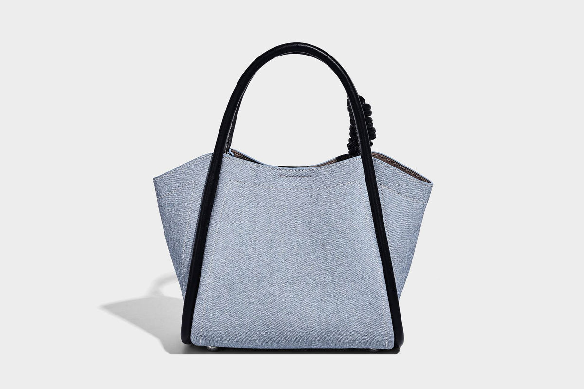 Light blue denim trapezoid tote bag with black leather-look trim, knotted top handles, detachable crossbody strap, front