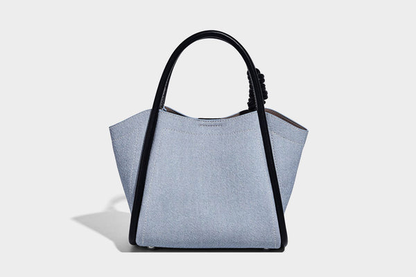 Light blue denim trapezoid tote bag with black leather-look trim, knotted top handles, detachable crossbody strap, front