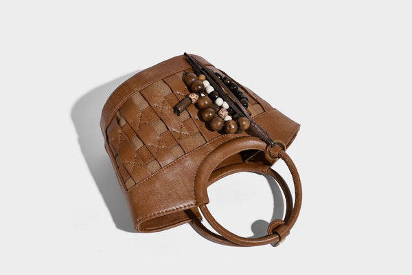 Small woven-panel bucket bag with circular ring handles, beaded tassel charm, and detachable crossbody strap in brown, detail