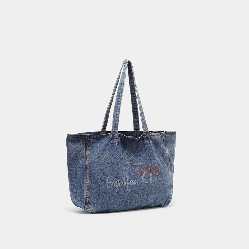 Large washed blue denim tote bag with handwritten-style graphic print, orange contrast stitching, and long fabric shoulder
