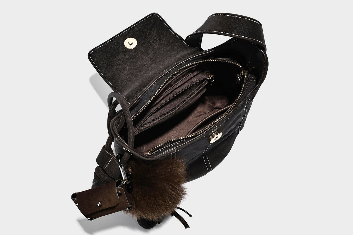 Small matte textured bucket crossbody bag with flap closure, gold-tone bar hardware, contrast stitching, and pompom tassel