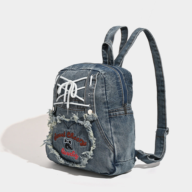 Washed denim blue backpack with white lace-up cord front panel, frayed heart-shaped embroidered exterior pocket, zip top