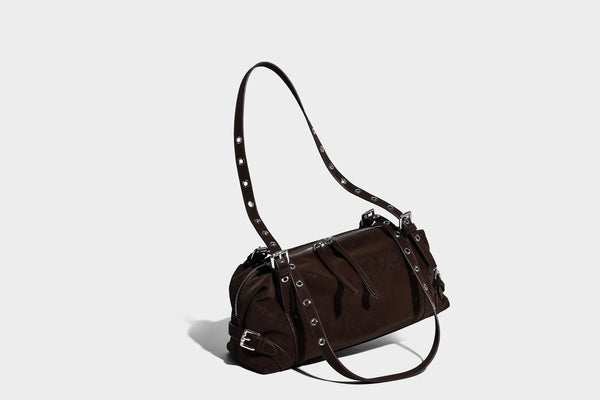 Weiche, zylinderförmige Schultertasche in Dunkelbraun mit silberfarbenen Riemen mit Ösen, Reißverschluss oben, seitlichen Schnallenbeschlägen und Quaste