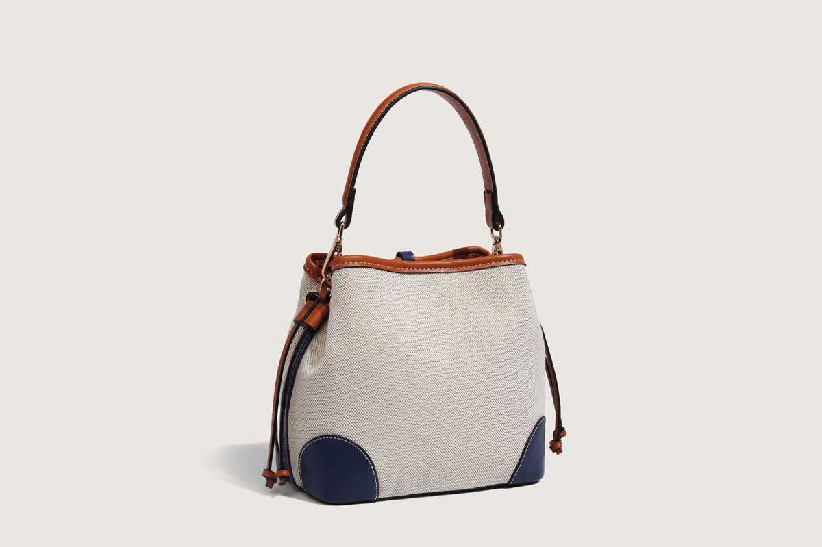 Cream woven-texture canvas bucket bag with navy accent panels, tan leather-look trim, gold-tone chain strap, and tassel