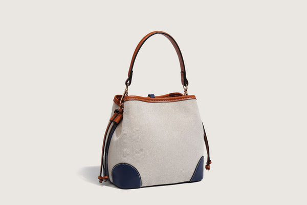 Cream woven-texture canvas bucket bag with navy accent panels, tan leather-look trim, gold-tone chain strap, and tassel