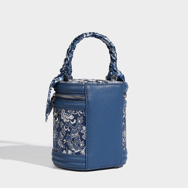 Small cylindrical barrel bag in blue-and-white floral jacquard with leather-look trim, scarf-wrapped top handle, silver-tone