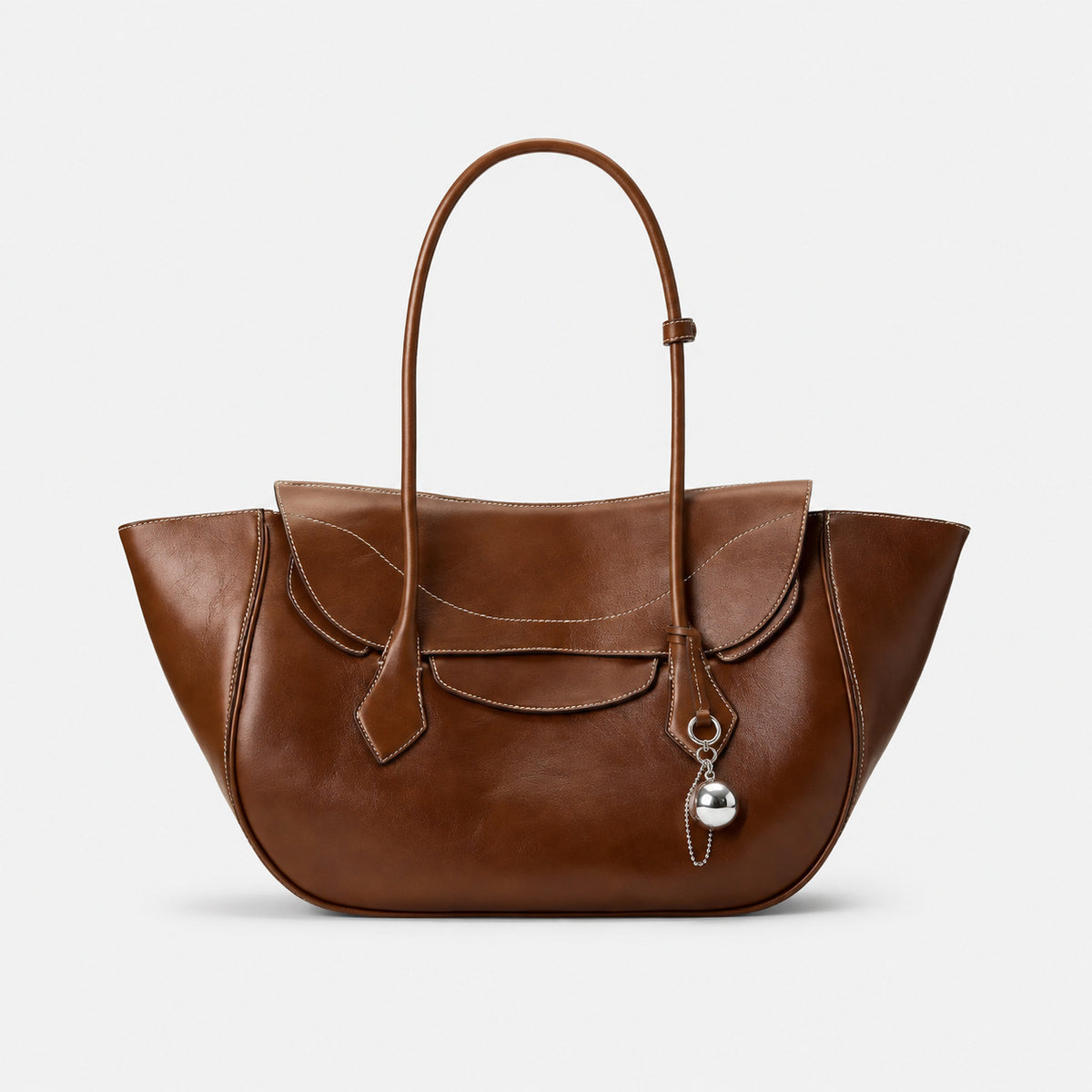 Large winged leather-look tote bag with single arched handle, stitched flap-front panel, and silver ball charm in cognac