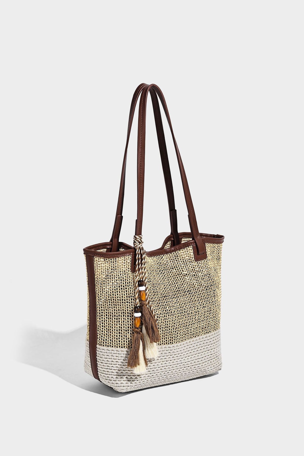 Woven beige and cream tote bag with brown leather-look trim, long double shoulder straps, and knotted tassel charm on front