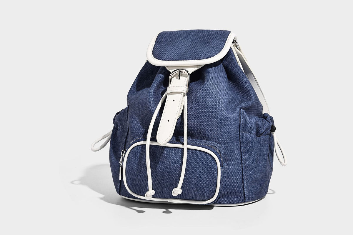 Compact denim drawstring backpack with white leather-look piping, flap-top buckle closure, front zip pocket, and silver-tone