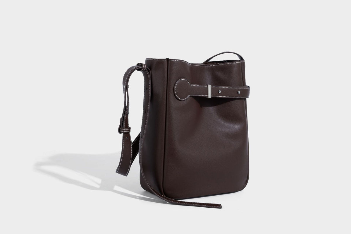 Structured dark brown leather-look bucket bag with white contrast stitching, front wraparound belt buckle strap, and long