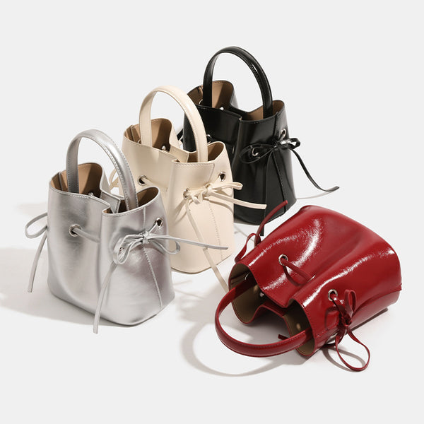 Compact glossy leather-look bucket bag with drawstring bow-tie closure, rounded top handle, and silver-tone grommet hardware