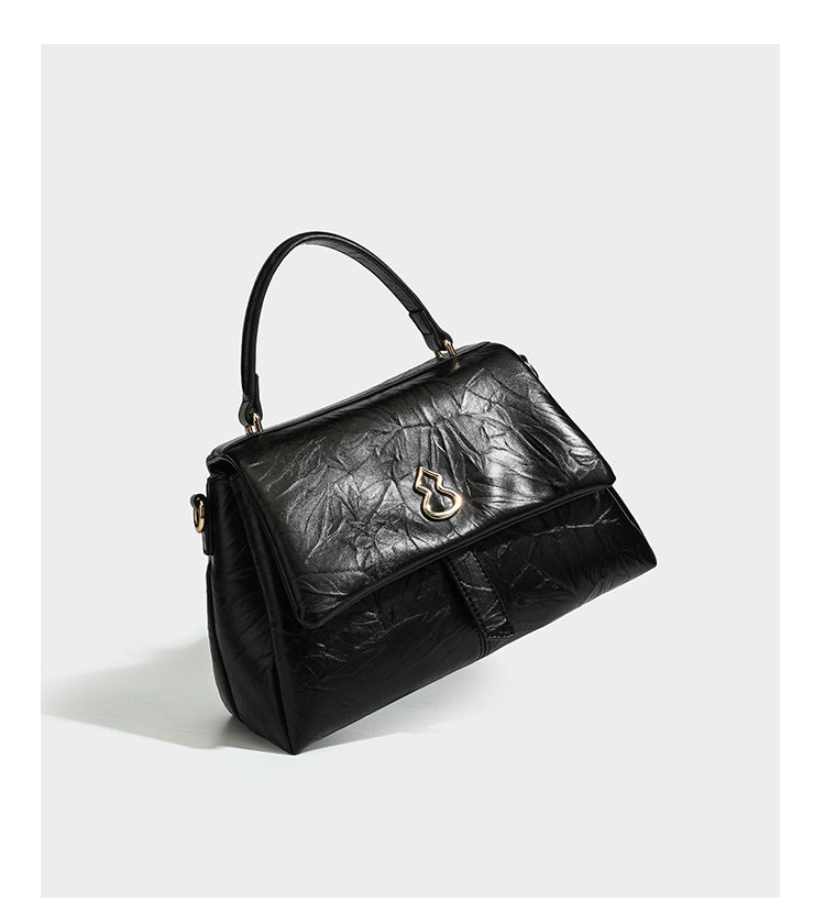 Structured black satchel bag with glossy crinkled leather-look finish, gold-tone emblem flap closure, slim top handle, and