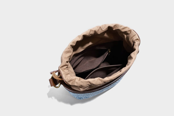Small light blue woven bucket bag with brown leather-look trim, braided top handle, brass-tone ring hardware, drawstring