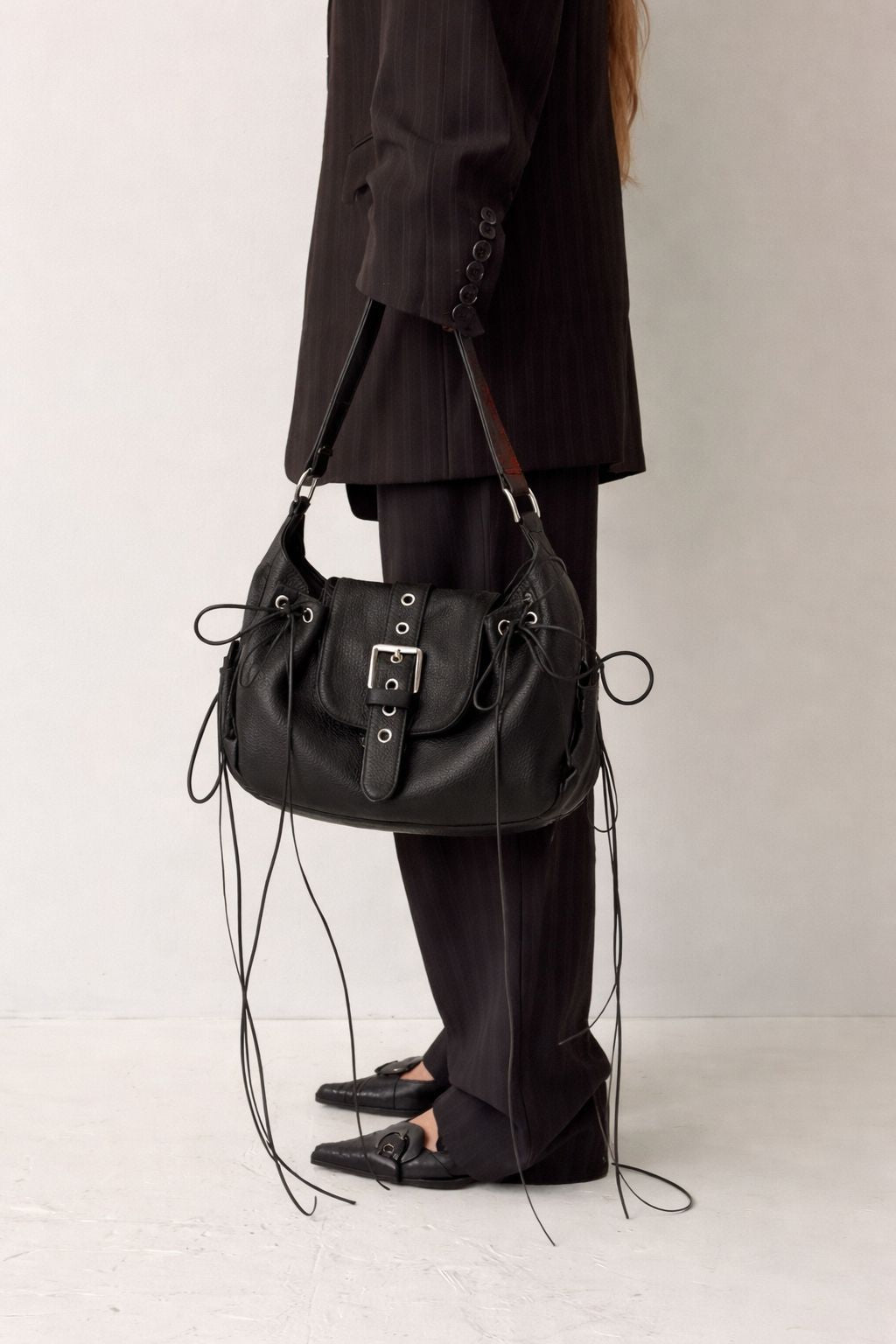 Schwarze Sattelschultertasche in genarbter Lederoptik mit geschnürten Seiteneinsätzen, Ösendetails und Klappe mit silberfarbener Schnalle, Detailansicht