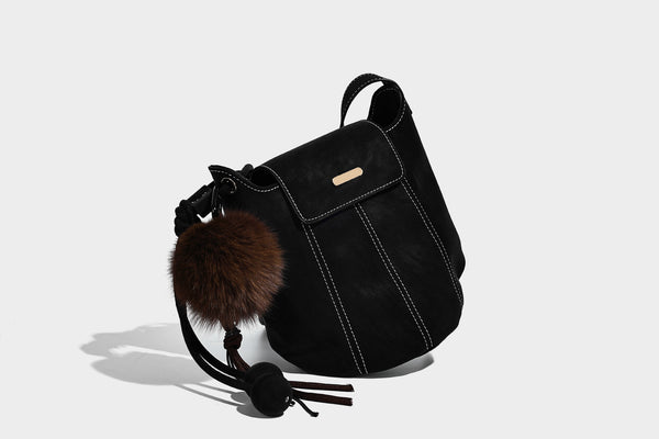 Small matte textured bucket crossbody bag with flap closure, gold-tone bar hardware, contrast stitching, and pompom tassel