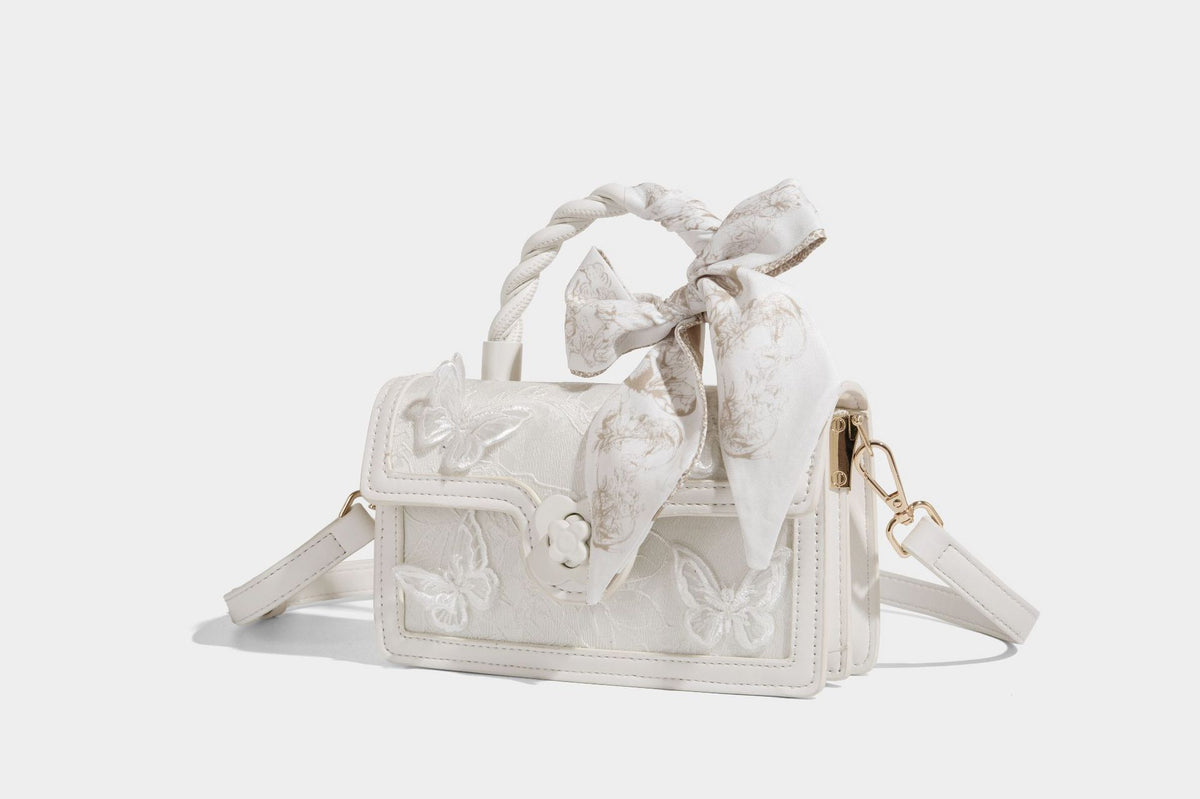 Small structured white flap bag with butterfly embroidery, flower-shaped turn-lock clasp, braided top handle with scarf bow