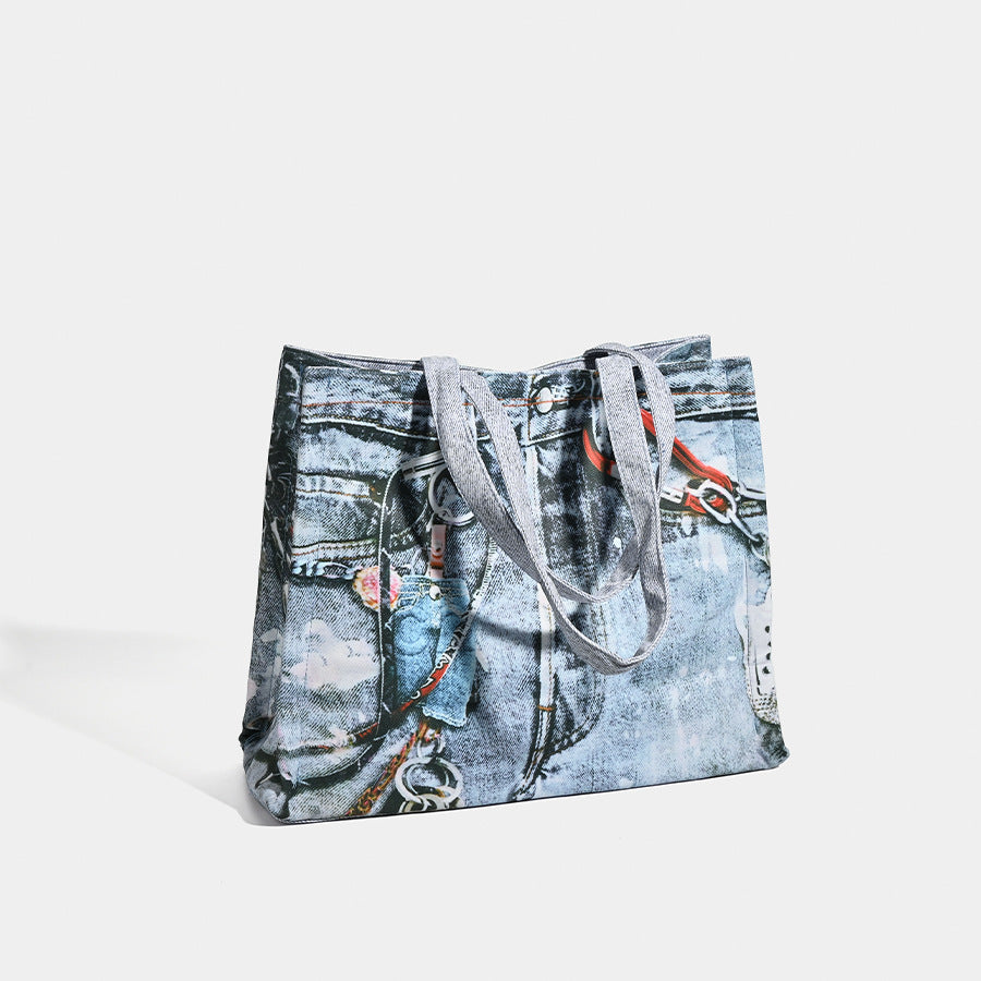 Large open-top tote bag with printed denim jeans graphic in faded blue-grey, dual flat shoulder straps and rivet hardware