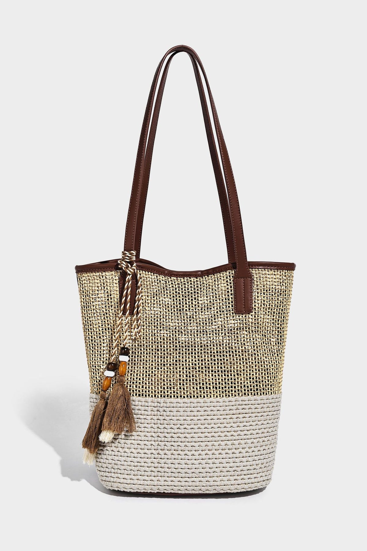 Woven straw-look tote bag in tan and cream with dark brown leather-look trim, long flat shoulder straps, and tassel bead